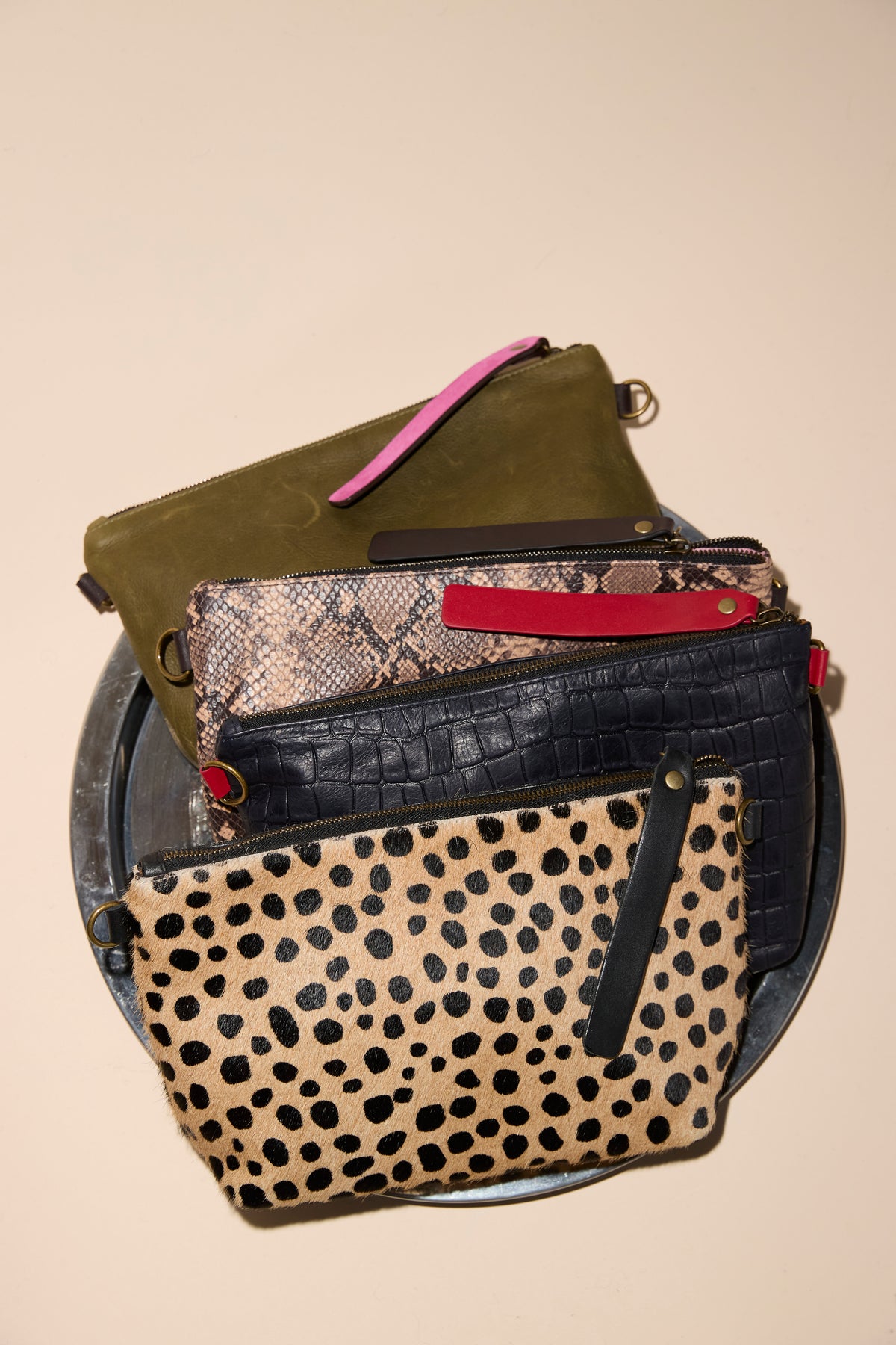 Four zip pouches stacked on a metal tray: olive leather with pink pull, snake-print with red pull, black croc-embossed, and spotted calf hair with black pull