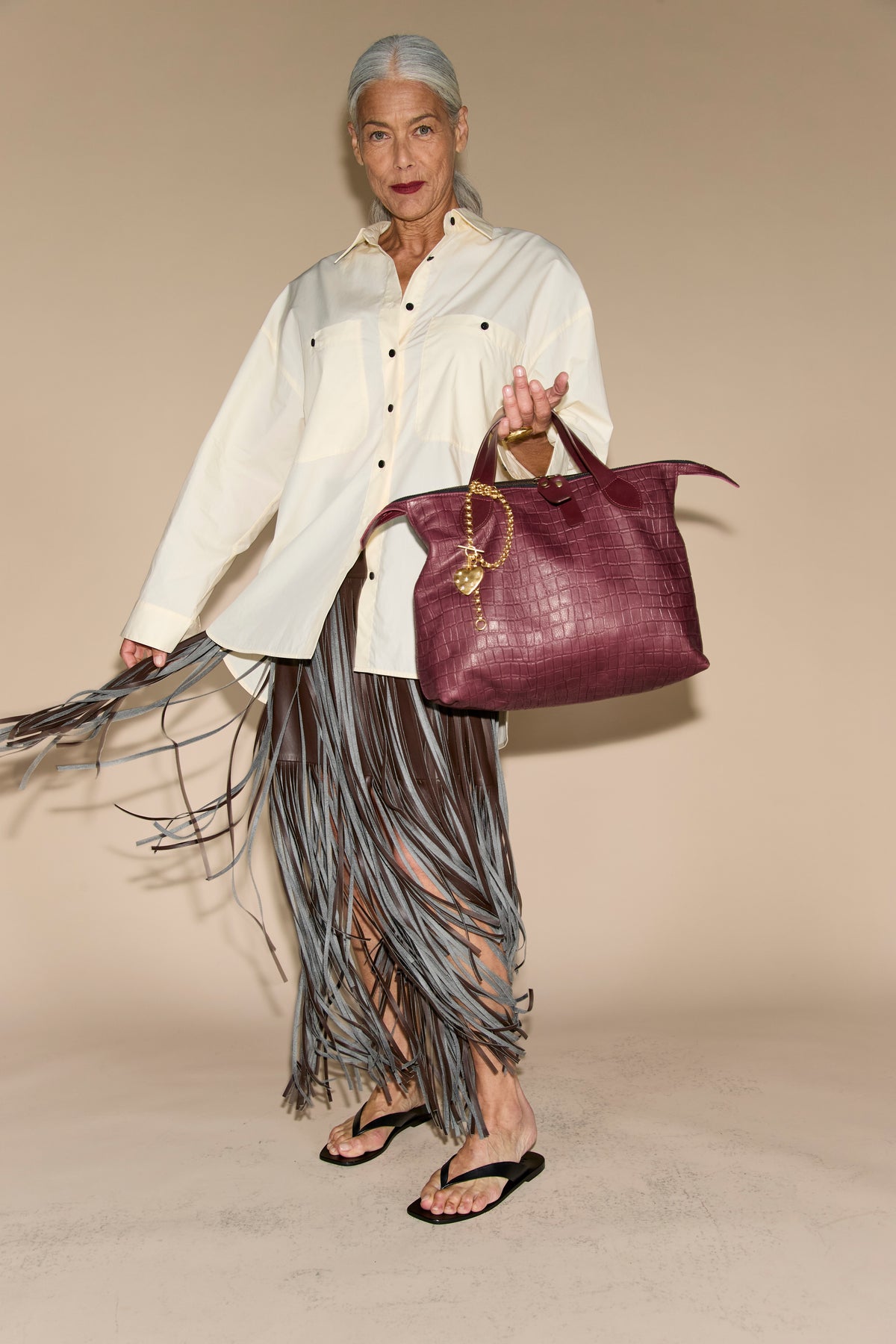Burgundy croc-embossed leather tote with dark leather handles and a gold heart charm on a chain, carried by hand by a standing model